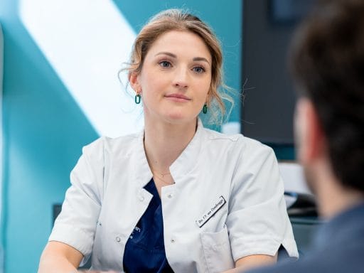 Vrouwelijke arts met blond haar draagt een witte jas en luistert aandachtig.