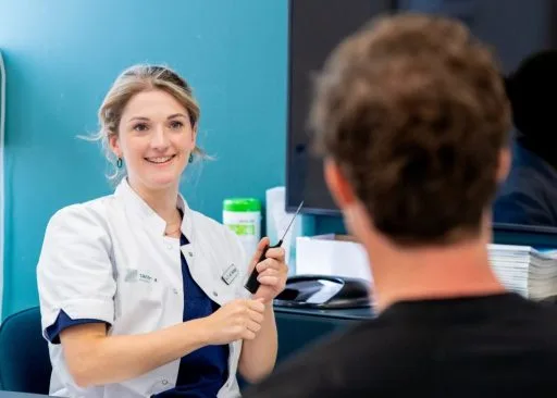 Vrolijke vrouwelijke arts legt iets uit aan een patiënt tijdens een consult over haartransplantatie, glimlachend in witte jas.