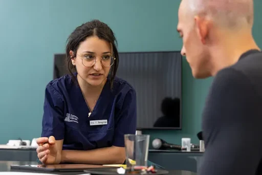 Vrouwelijke arts in blauwe scrubs met naamkaartje bespreekt een haartransplantatie met een kale man tijdens een consult.