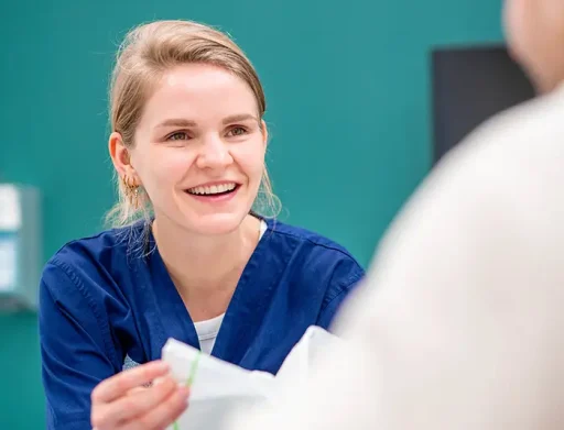 Lachende verpleegkundige in blauwe scrubs geeft nazorginstructies of materialen aan een patiënt na een haartransplantatie.