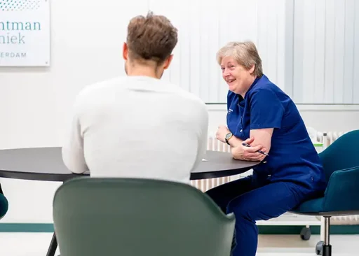 Verpleegkundige in blauwe scrubs in gesprek met mannelijke patiënt tijdens consult over haartransplantatie.