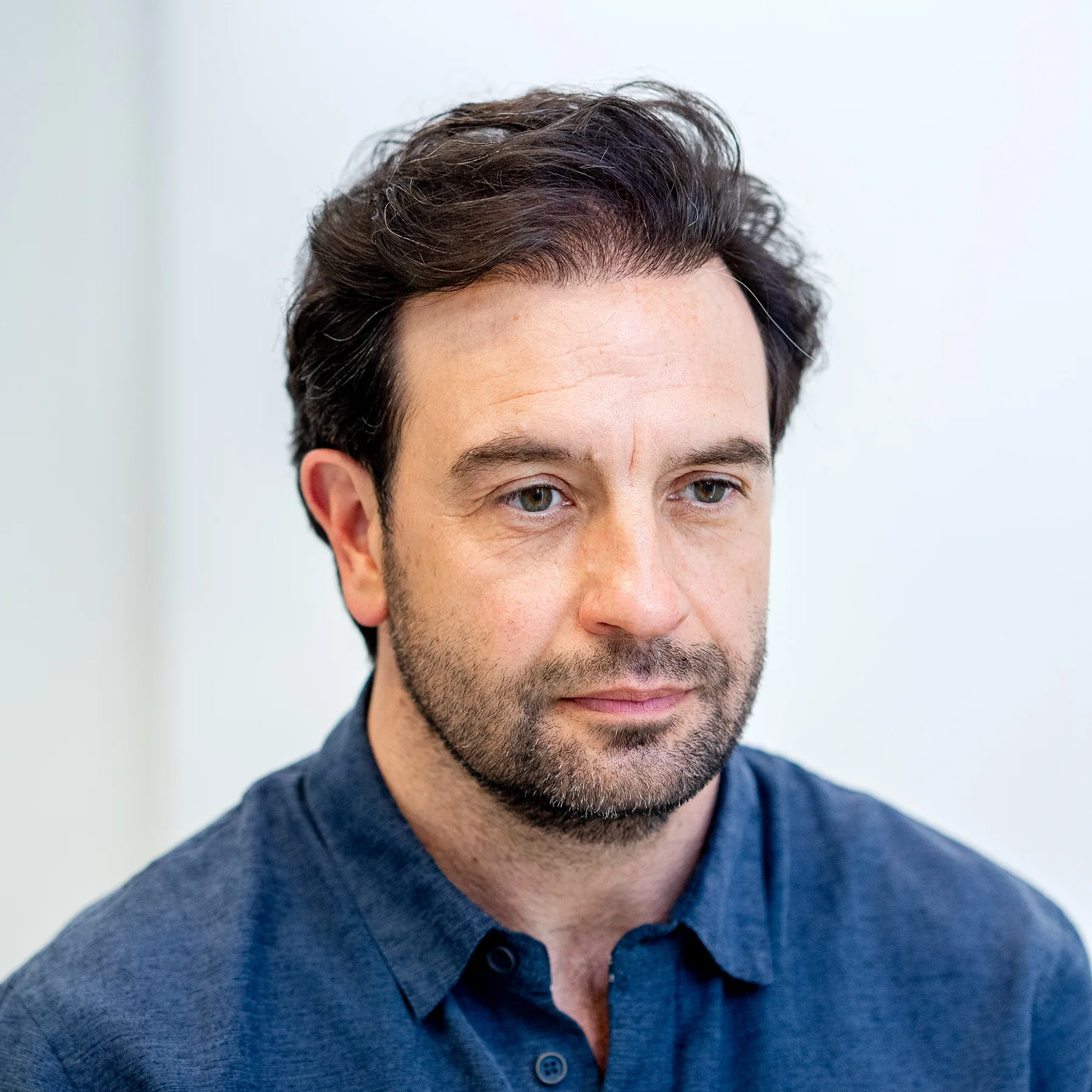 Man with dark curly hair and light beard looks serious in a dark blue shirt.