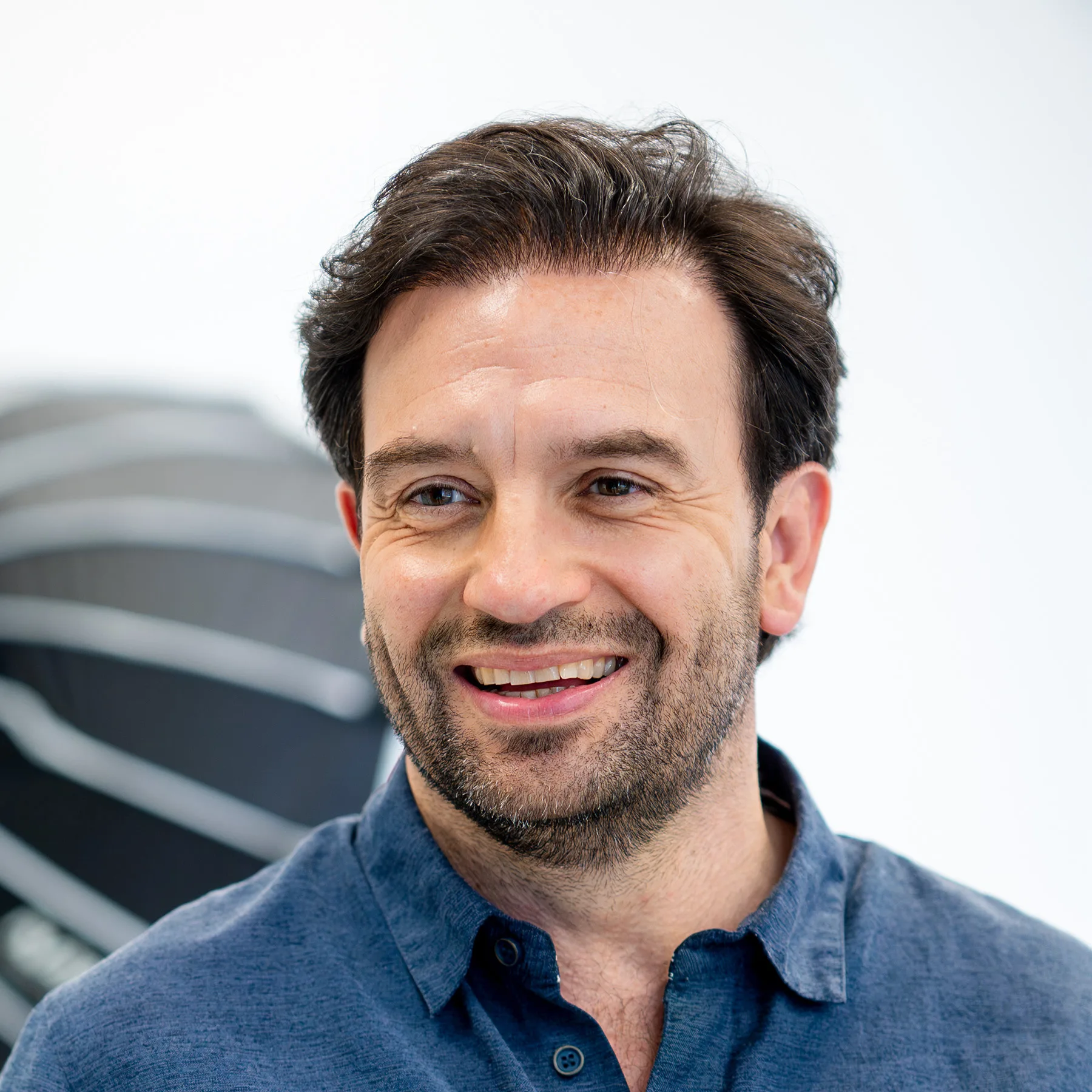 Man with dark hair and stubble smiles in a blue shirt.