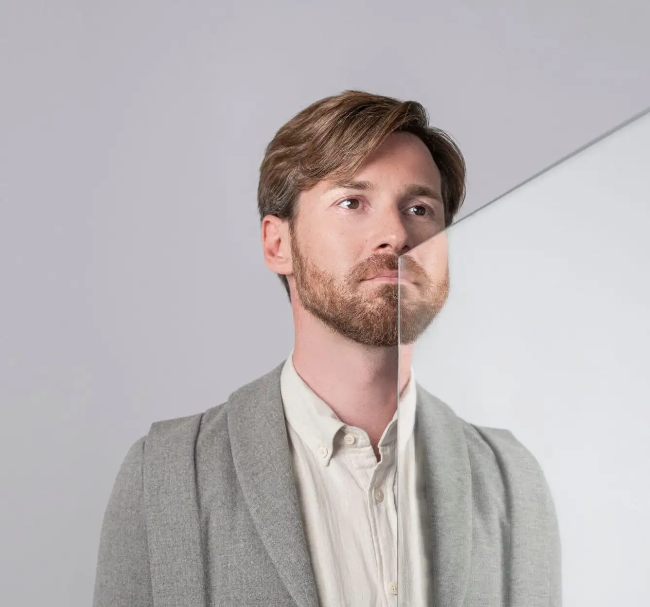 Man with full beard and neat clothing, with a glass plate reflecting part of his face, symbolizing beard transplant results.